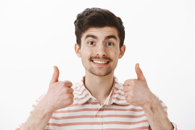 Anímate hombre, todo genial. Retrato de chico caucásico amistoso positivo con bigote, levantando los pulgares hacia arriba y sonriendo ampliamente, aprobando un nuevo concepto o idea de amigo, siendo alegre y satisfecho