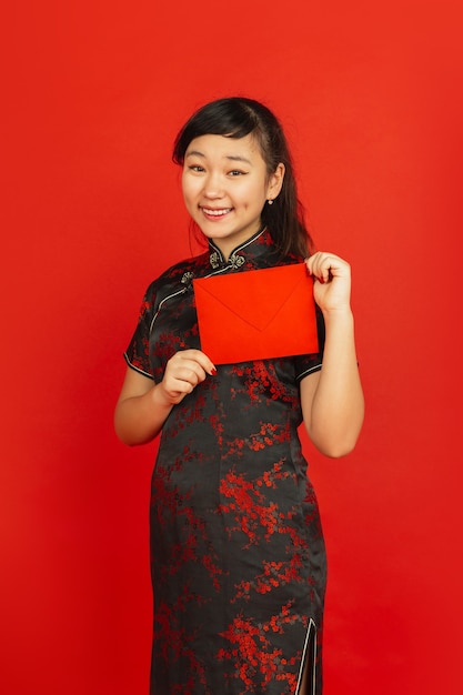 Foto gratuita año nuevo chino 2020. retrato de joven asiática aislado sobre fondo rojo. modelo femenino en ropa tradicional se ve feliz, sonriendo y sosteniendo un sobre rojo. celebración, fiesta, emociones.