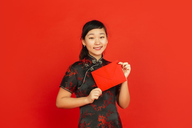 Año nuevo chino 2020. Retrato de joven asiática aislado sobre fondo rojo. Modelo femenino en ropa tradicional se ve feliz, sonriendo y sosteniendo un sobre rojo. Celebración, fiesta, emociones.