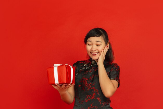 Año nuevo chino 2020. Retrato de joven asiática aislado sobre fondo rojo. Modelo femenino en ropa tradicional se ve feliz, sonriente y sorprendida por caja de regalo. Celebración, fiesta, emociones.