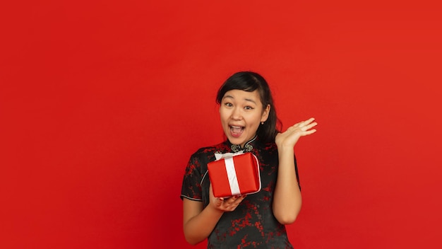 Año Nuevo Chino. Retrato de niña asiática aislado sobre fondo rojo. Modelo femenino en ropa tradicional se ve feliz, sonriente y sorprendida por caja de regalo. Celebración, fiesta, emociones.