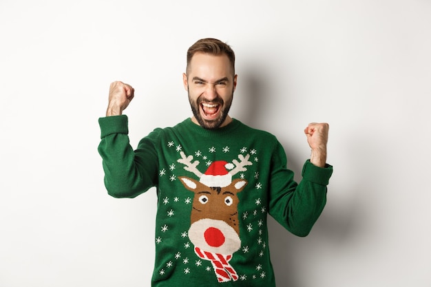 Año nuevo, fiestas y celebración. Hombre barbudo emocionado en suéter de Navidad, haciendo puños y gritando de alegría, regocijo y triunfo, fondo blanco.