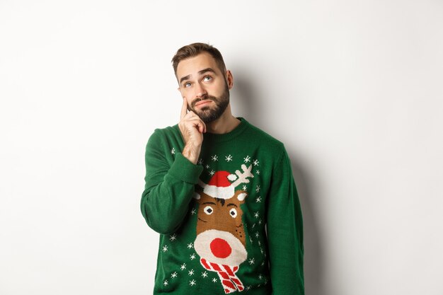 Año nuevo, fiestas y celebración. Triste joven con barba, vestido con suéter verde, mostrando una lágrima en su rostro y mirando molesto, de pie sobre fondo blanco.