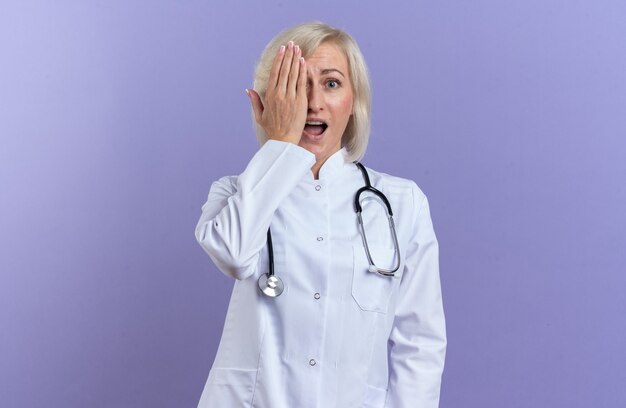 Ansiosa doctora eslava adulta en bata médica con estetoscopio que cubre el ojo con la mano aislada sobre fondo púrpura con espacio de copia