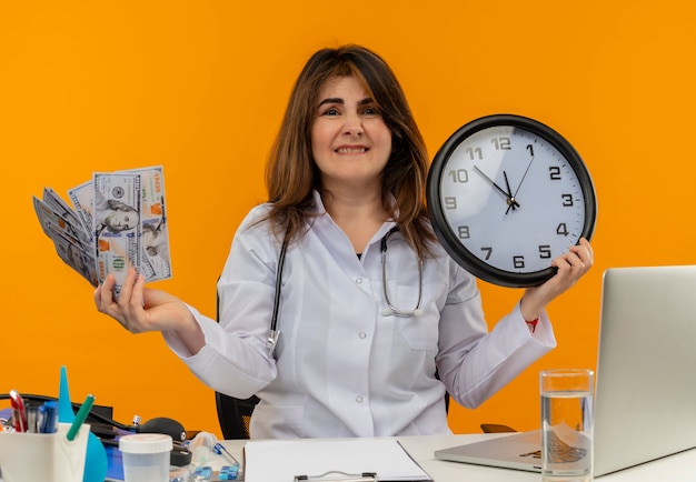 Ansiosa doctora de mediana edad con bata médica y estetoscopio sentado en el escritorio con portapapeles de herramientas médicas y computadora portátil con reloj y dinero mordiendo el labio aislado