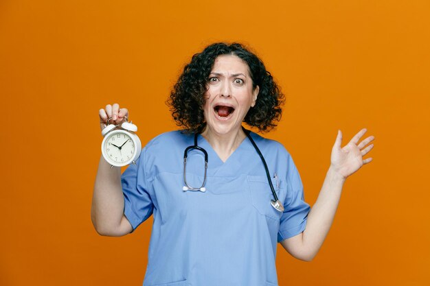 Ansiosa doctora de mediana edad con uniforme y estetoscopio alrededor del cuello mirando la cámara que muestra el despertador y la mano vacía aislada en el fondo naranja