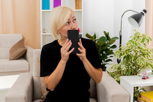 Ansiosa hermosa mujer rubia sentada en un sillón sosteniendo el teléfono y mirando al lado