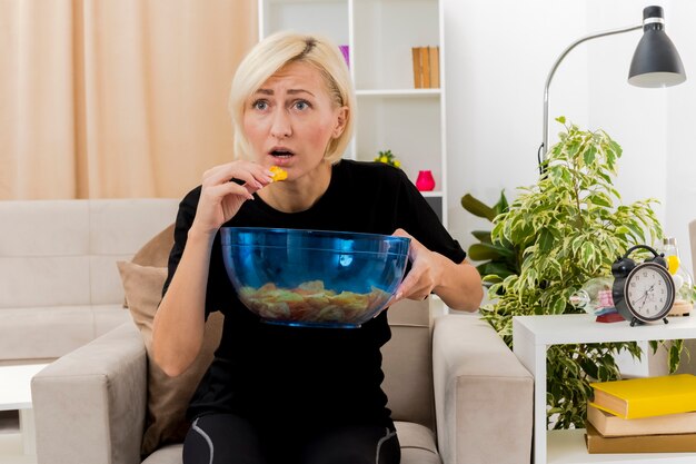 Ansiosa hermosa mujer rubia se sienta en el sillón sosteniendo y comiendo un tazón de patatas fritas