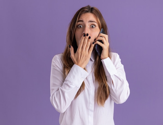 Ansiosa joven bastante caucásica pone la mano en la boca y habla por teléfono aislado en la pared púrpura con espacio de copia