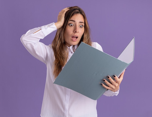 Ansiosa joven bastante caucásica pone la mano en la cabeza sosteniendo y mirando la carpeta de archivos en púrpura