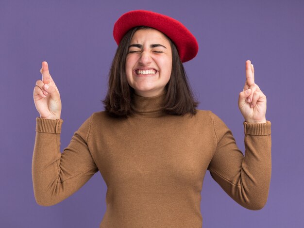 Ansiosa joven bastante caucásica con sombrero de boina se encuentra con los brazos cruzados aislados en la pared púrpura con espacio de copia