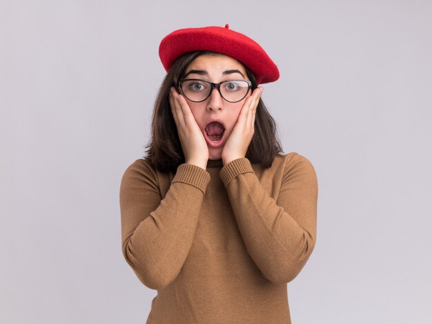 Ansiosa joven bastante caucásica con sombrero de boina y gafas ópticas pone las manos en la cara aislada en la pared blanca con espacio de copia