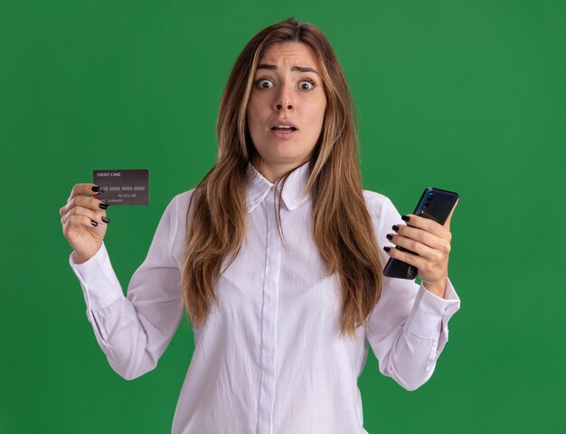 Ansiosa joven bastante caucásica tiene tarjeta de crédito y teléfono aislado en la pared verde con espacio de copia