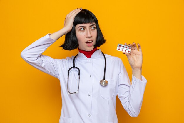 Ansiosa joven bastante caucásica en uniforme médico con estetoscopio poniendo la mano sobre su cabeza y sosteniendo el paquete de pastillas