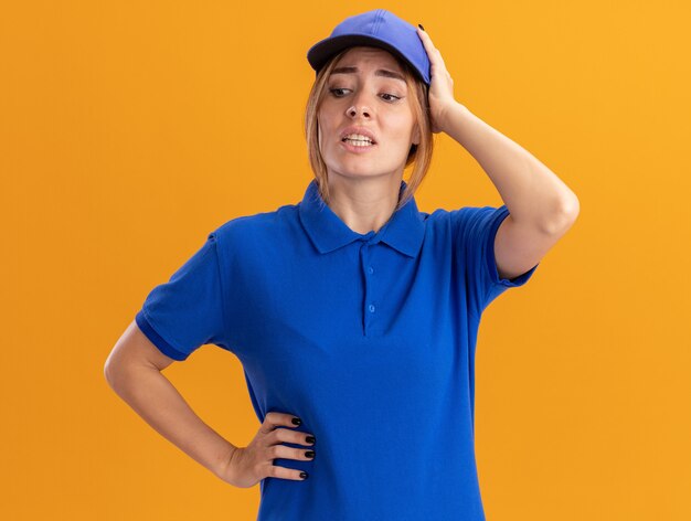 Ansiosa joven bonita repartidora en uniforme pone la mano en la cabeza y mira hacia abajo en naranja