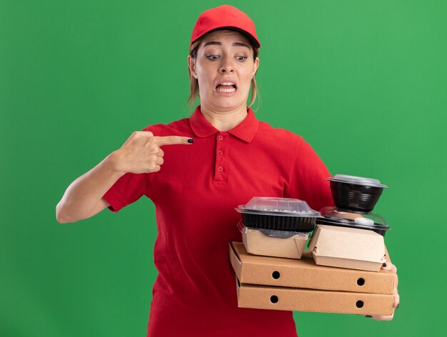 Ansiosa joven bonita repartidora en uniforme sostiene y apunta a paquetes de alimentos de papel y contenedores en cajas de pizza en verde