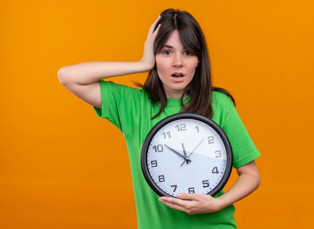 Ansiosa joven caucásica en camisa verde sostiene el reloj y pone la mano en la cabeza sobre fondo naranja aislado