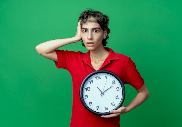 Ansiosa joven caucásica con corte de pelo pixie sosteniendo el reloj poniendo la mano en la cabeza aislada sobre fondo verde