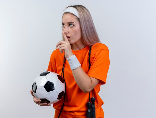 Ansiosa joven caucásica deportiva con tirantes y con saltar la cuerda alrededor del cuello con diadema y muñequeras sostiene la bola y gestos signo de silencio mirando de lado en la pared blanca