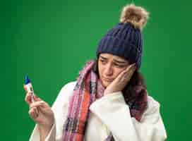 Foto gratuita ansiosa joven caucásica enferma vistiendo bata gorro de invierno y bufanda sosteniendo y mirando el termómetro manteniendo la mano en la cara aislada en la pared verde