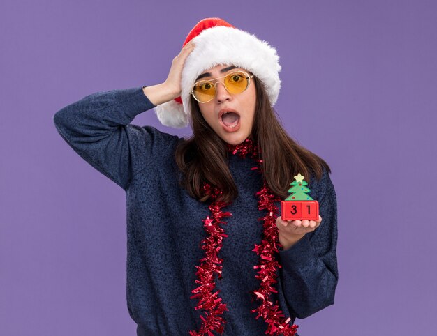 Ansiosa joven caucásica en gafas de sol con gorro de Papá Noel y guirnalda alrededor del cuello pone la mano en la cabeza y sostiene el adorno del árbol de Navidad aislado en la pared púrpura con espacio de copia