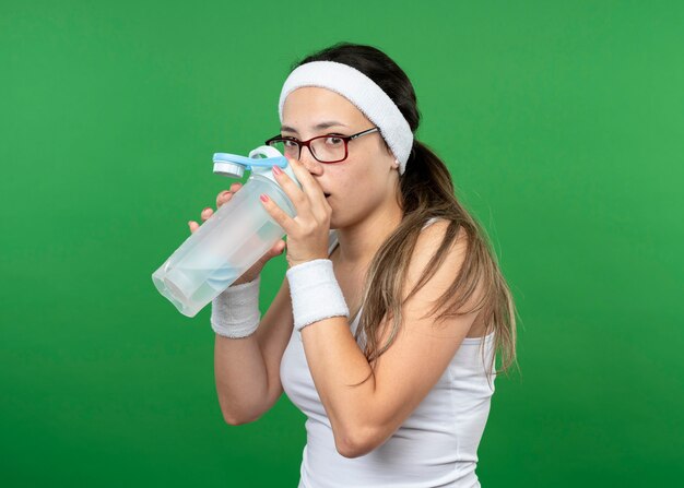 Ansiosa joven deportiva en gafas ópticas con diadema y muñequeras bebe de la botella de agua