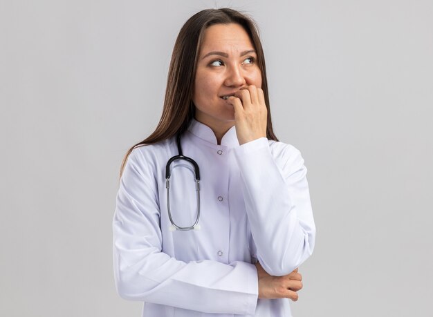 Ansiosa joven doctora asiática vistiendo bata médica y estetoscopio mordiendo los dedos mirando al lado aislado en la pared blanca con espacio de copia