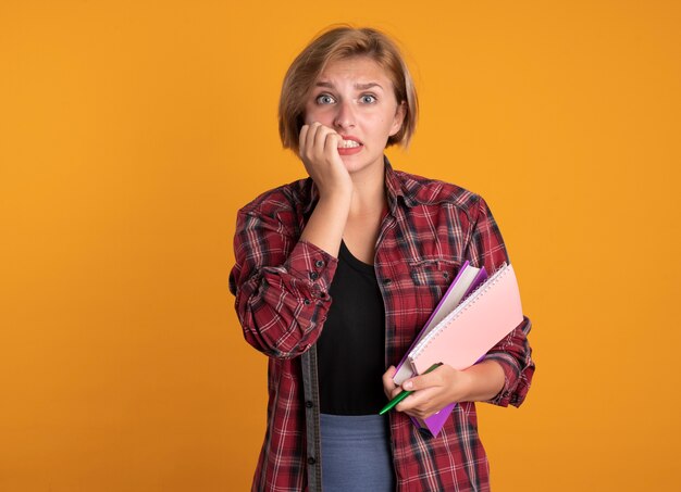 Ansiosa joven estudiante eslava se muerde las uñas y sostiene el cuaderno y el bolígrafo