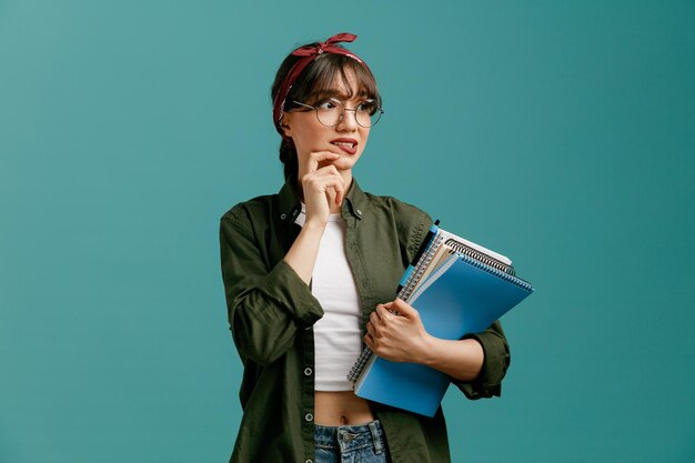 Ansiosa joven estudiante con gafas de bandana sosteniendo grandes blocs de notas con bolígrafo mirando el lado tocando la barbilla mordiendo el labio aislado en el fondo azul