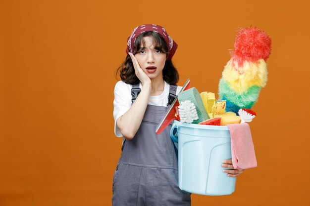 Ansiosa joven limpiadora con uniforme y pañuelo sosteniendo un balde de herramientas de limpieza manteniendo la mano en la cara mirando la cámara aislada en el fondo naranja
