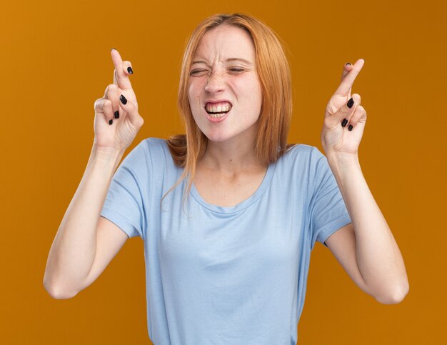 Ansiosa joven pelirroja pelirroja con pecas se encuentra con los ojos cerrados cruzando los dedos en naranja