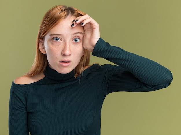 Foto gratuita ansiosa joven pelirroja pelirroja con pecas pone la mano en la frente aislada en la pared verde oliva con espacio de copia