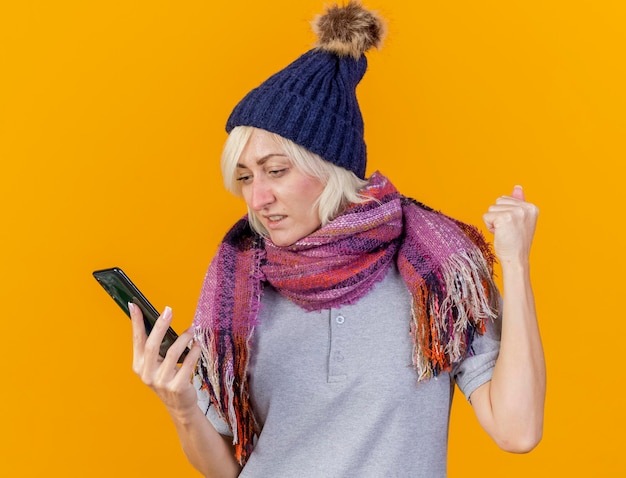 Ansiosa joven rubia enferma eslava vistiendo gorro y bufanda de invierno mantiene el puño en alto