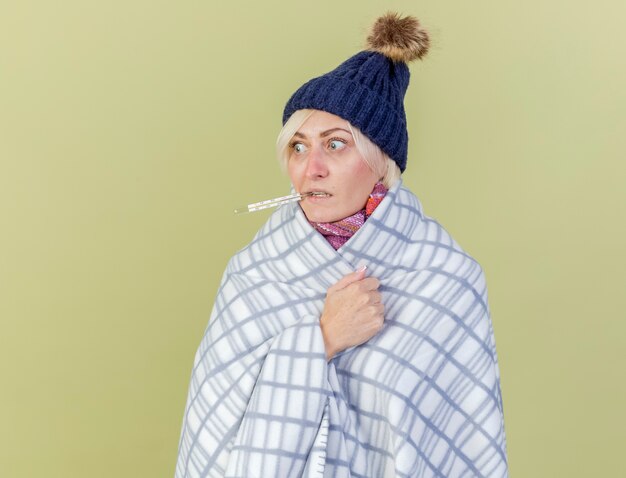 Ansiosa joven rubia enferma mujer eslava con gorro de invierno y bufanda envuelta en cuadros tiene termómetro con dientes mirando al lado aislado en la pared verde oliva con espacio de copia