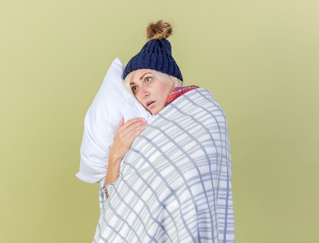 Ansiosa joven rubia enferma vistiendo gorro de invierno y bufanda envuelta en cuadros abrazos almohada mirando al lado aislado en la pared verde oliva