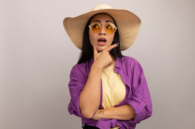 Ansiosa mujer bonita morena en gafas de sol con sombrero de playa sostiene la barbilla y mira al lado aislado en la pared blanca