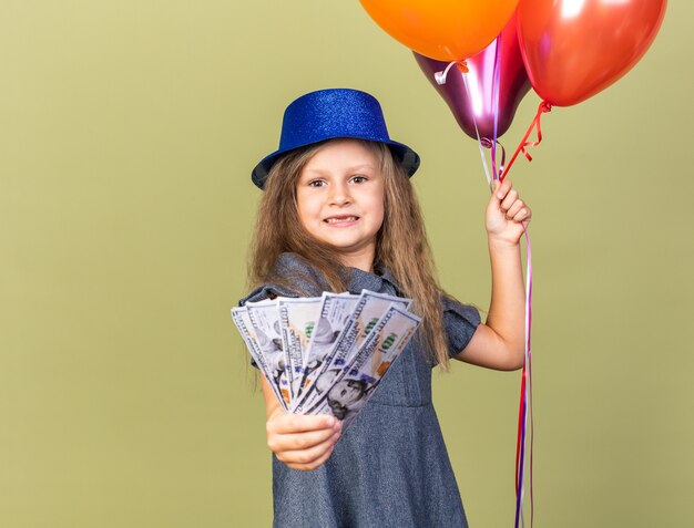 Ansiosa niña rubia con gorro de fiesta azul sosteniendo globos de helio y dinero aislado en la pared verde oliva con espacio de copia