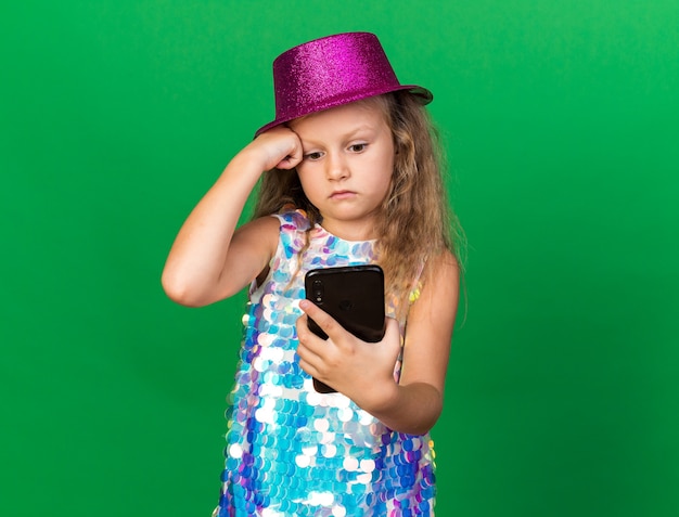 Ansiosa niña rubia con gorro de fiesta púrpura sosteniendo y mirando el teléfono aislado en la pared verde con espacio de copia