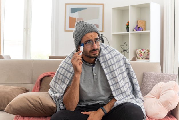 Ansioso enfermo joven en gafas ópticas envuelto en cuadros con gorro de invierno sosteniendo el termómetro mirando al lado sentado en el sofá en la sala de estar