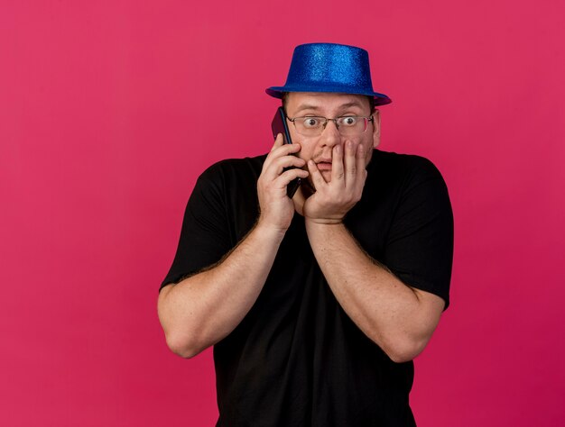 Ansioso hombre eslavo adulto en gafas ópticas con gorro de fiesta azul pone la mano en la boca hablando por teléfono