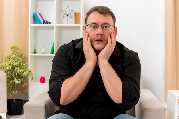 Ansioso hombre eslavo adulto con gafas ópticas se sienta en un sillón poniendo las manos en la cara dentro de la sala de estar diseñada