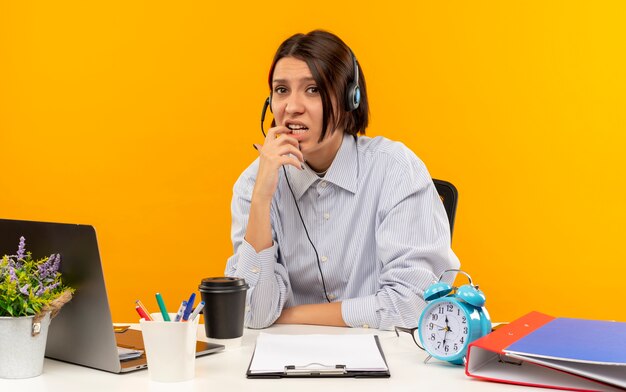 Ansioso joven call center chica con auriculares sentado en el escritorio poniendo el dedo en el labio aislado en naranja