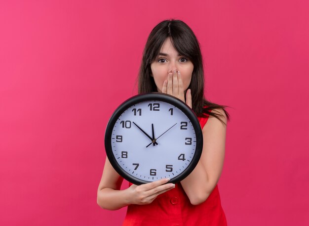 Ansioso joven caucásica sosteniendo el reloj y sosteniendo la boca mirando a la cámara sobre fondo rosa aislado