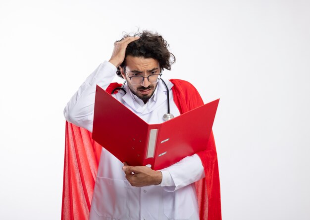 Ansioso joven caucásico con gafas ópticas con uniforme médico con manto rojo y con estetoscopio alrededor del cuello pone la mano en la cabeza sosteniendo y mirando la carpeta de archivos con espacio de copia