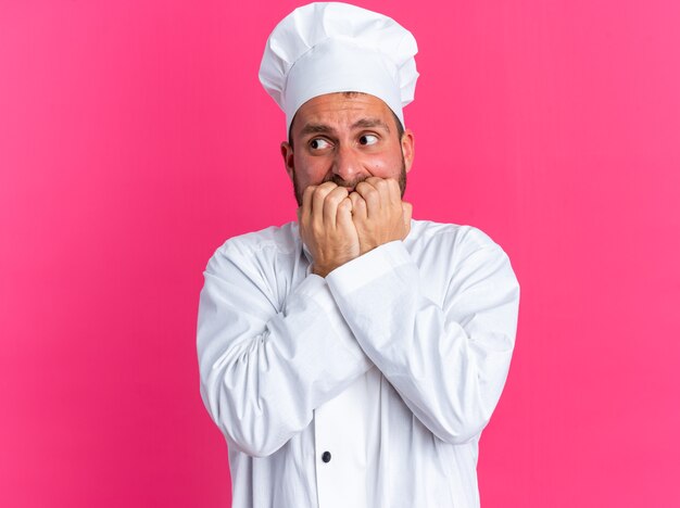 Ansioso joven cocinero de sexo masculino caucásico en uniforme de chef y gorra mirando al lado mordiendo los dedos