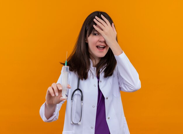 Ansioso joven doctora en bata médica con estetoscopio sostiene la jeringa y sostiene la cabeza sobre fondo naranja aislado con espacio de copia