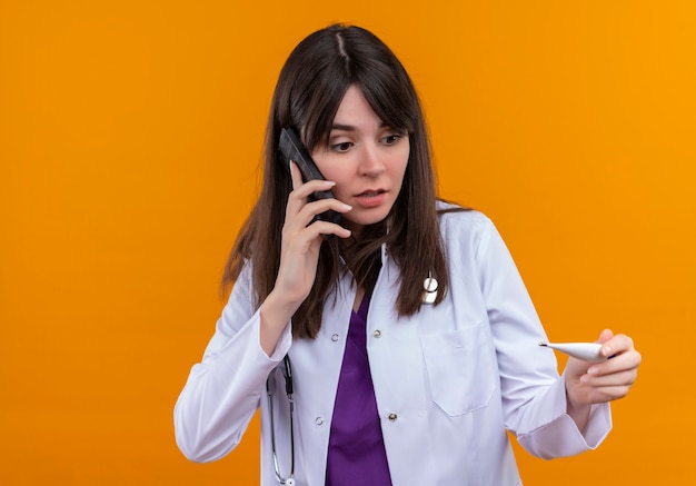 Ansioso joven doctora en bata médica con estetoscopio sostiene termómetro y habla por teléfono sobre fondo naranja aislado con espacio de copia