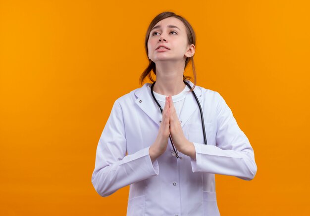 Ansioso joven doctora vistiendo bata médica y estetoscopio haciendo gesto de oración en un espacio naranja aislado con espacio de copia