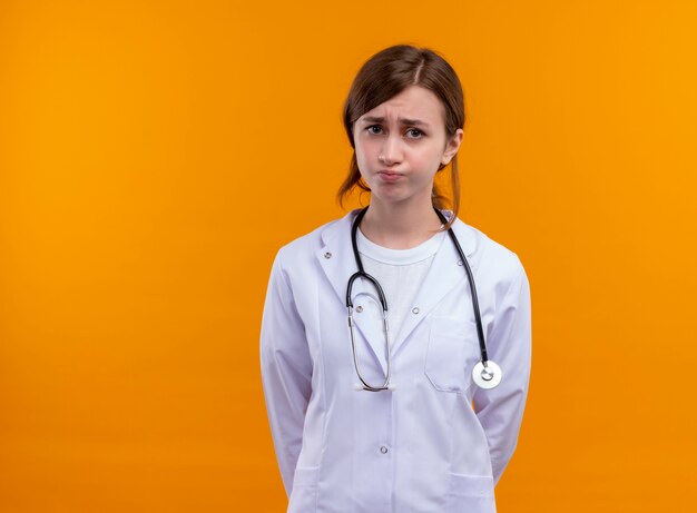 Ansioso joven doctora vistiendo bata médica y estetoscopio de pie en un espacio naranja aislado con espacio de copia