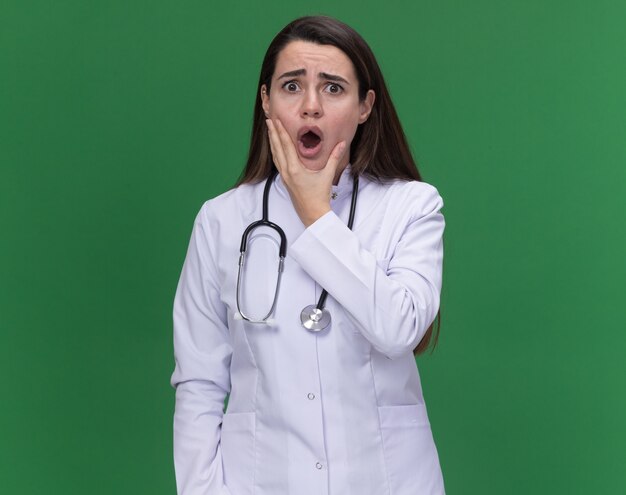 Ansioso joven doctora vistiendo bata médica con estetoscopio sostiene la barbilla y mira a la cámara en verde